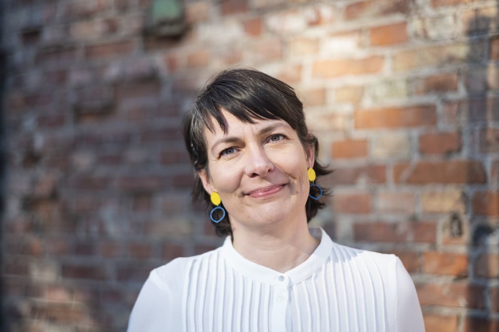 Kadi Viik, tumeda lühikese soengu ja tukaga, kannab valget plisseeritud pluusi ning erkkollaseid ringikujulisi kõrvarõngaid . Taustal on hägustatult näha punakaspruun päikeseline tellismüür. Portreefoto on tehtud päevavalguses, Viik vaatab kaamerasse sooja naeratusega.